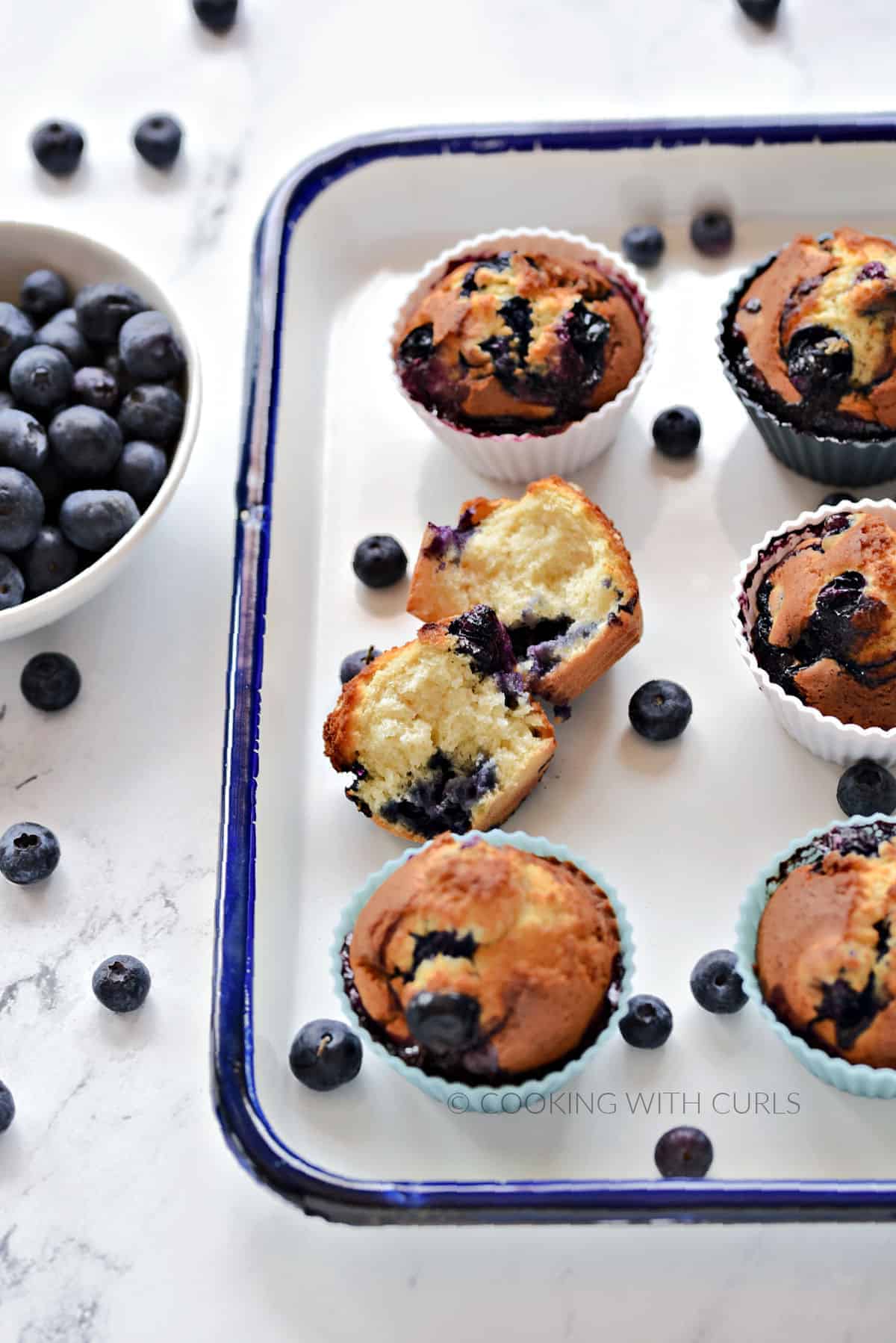 One split and five whole blueberry muffins on a white tray with a bowl of fresh blueberries off to the side. 