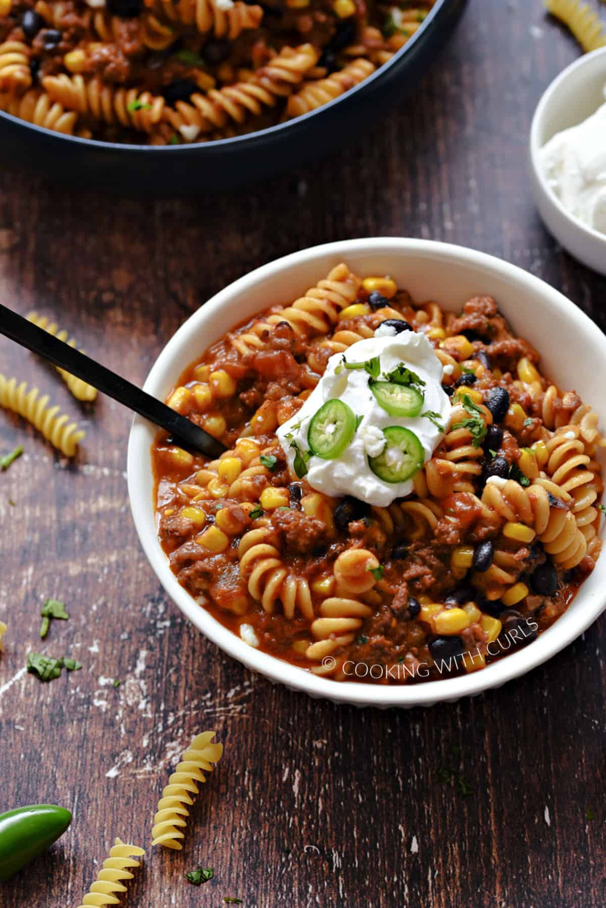 Taco pasta with beans and corn in a white bowl topped with sour cream and sliced jalapeno.