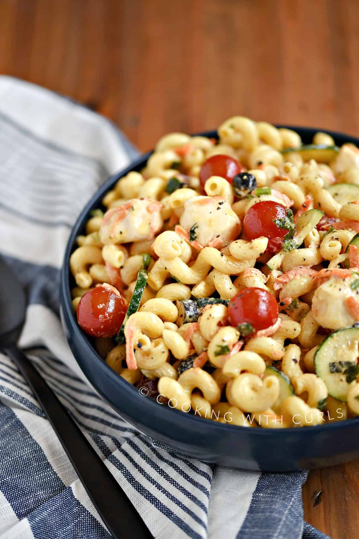 Spiral pasta, mozzarella cheese, cherry tomatoes, zucchini and black olives mixed together in a blue bowl.
