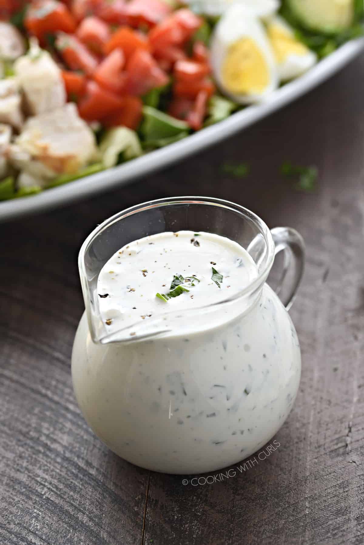 Homemade ranch dressing in a small glass pitcher with a classic cobb salad in the background.