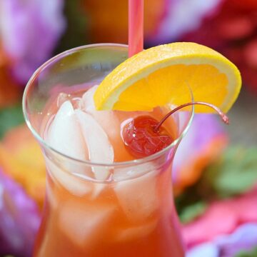 Bright red drink in a hurricane glass filled with ice and garnished with an orange wedge and cherry.