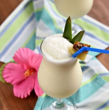 Creamy white drink in a poco grande glass with a pineapple wedge, blue straw, and two pineapple leaves.