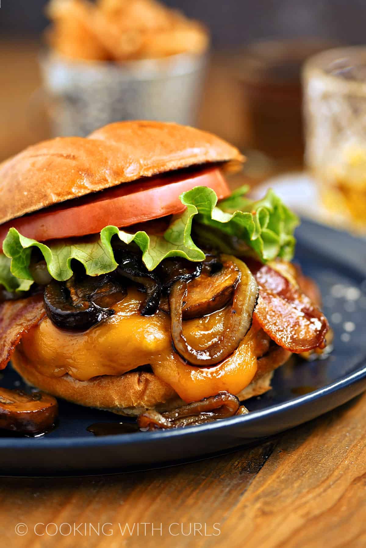 Burger topped with melted cheddar cheese, onions, mushrooms and glaze with a metal bucket of fries on the side.