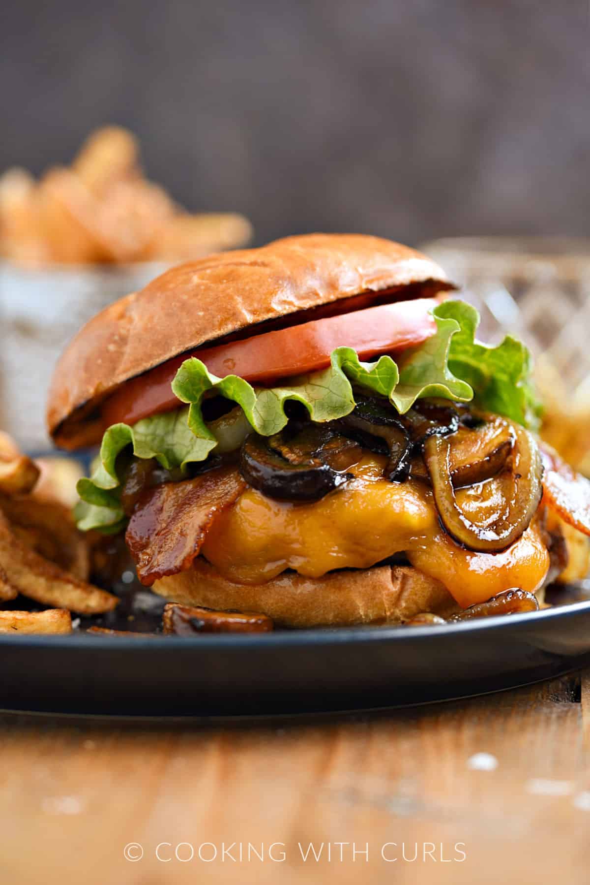 A burger topped with melted cheddar cheese, caramelized onions and mushrooms, lettuce , tomato, and whiskey glaze with fries on the side.
