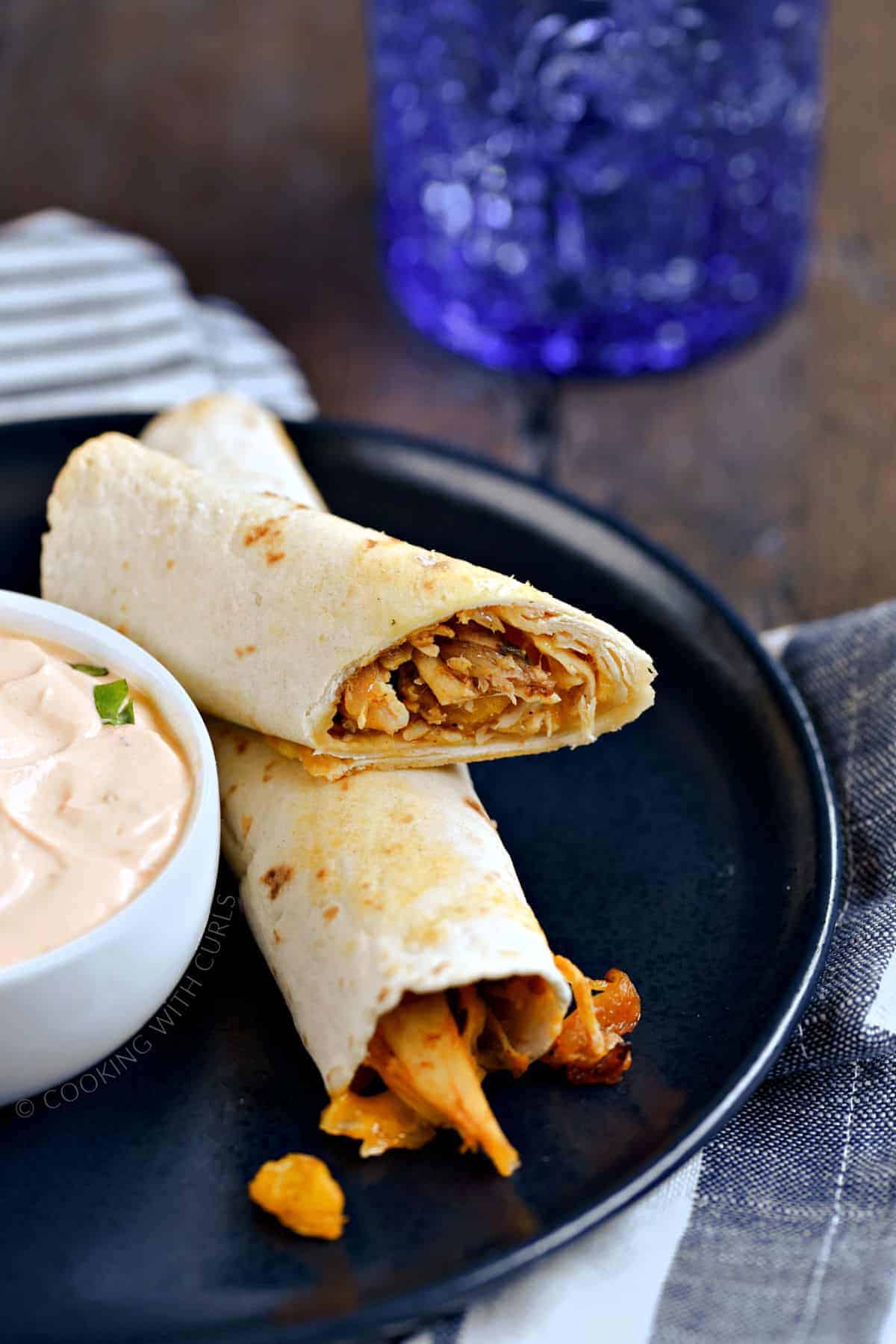 A full and one half peach chicken taquitos on a blue plate with a bowl creamy adobo sauce and a blue glass in the background. 
