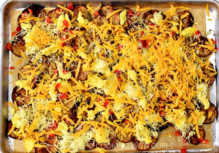 Baked potato slices topped with chopped red bell pepper, scrambled eggs, and shredded cheese on a baking sheet. 