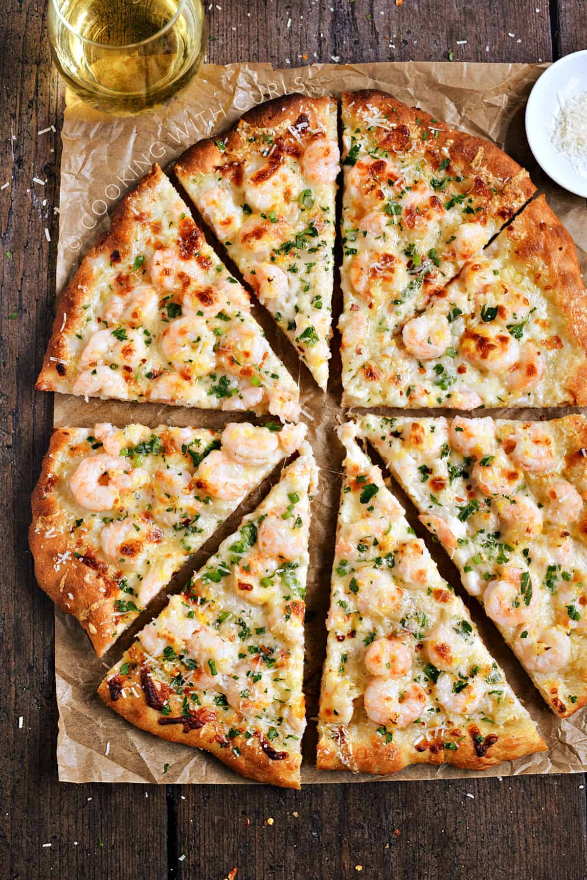 A sliced, shrimp pizza on a sheet of parchment paper with a glass of white wine in the left corner and bowl of Parmesan on the right.