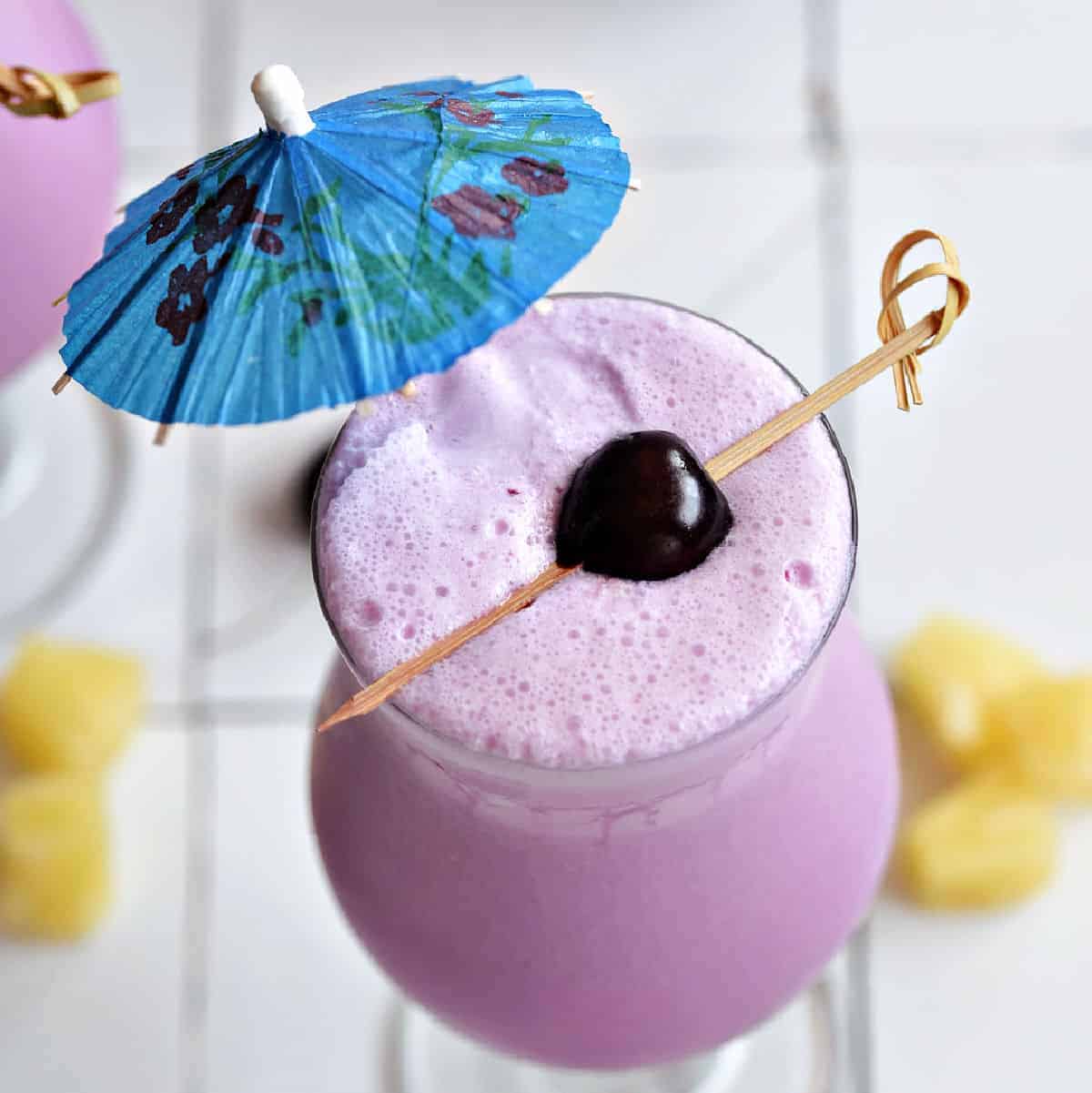 Frozen pink cocktail with blue umbrella and fresh cherry on a bamboo stick.