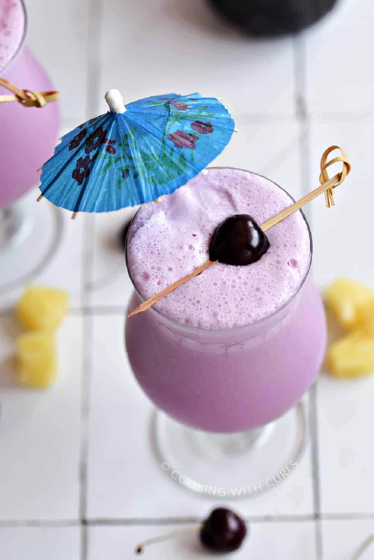 Frozen pink cocktail with a blue umbrella and fresh cherry on a bamboo stick.