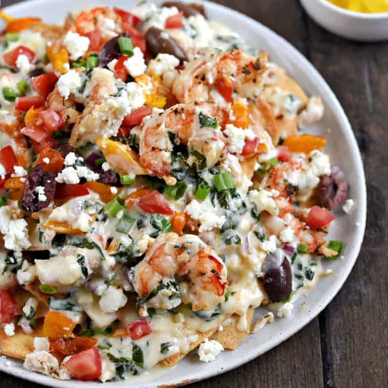 A large plate topped with shrimp, crab, diced tomatoes, olives, orange peppers, green onions, pita chips, and spinach cheese sauce.