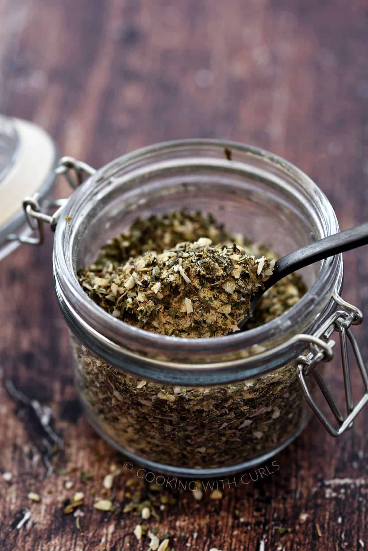 A small glass jar filled with Greek herbs and spices.