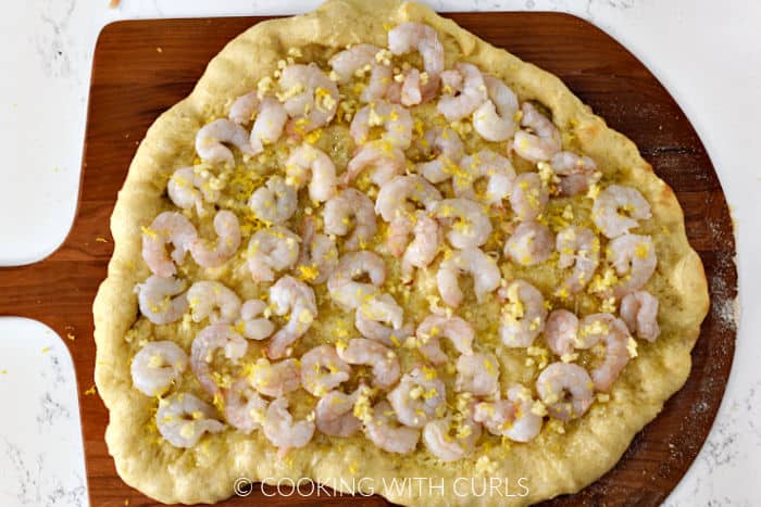 Pizza crust topped with raw shrimp, minced garlic, and lemon zest on a wooden pizza peel. 