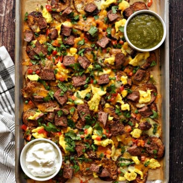 Baked sliced potatoes topped with chopped red pepper and scrambled eggs on a baking sheet with bowls of green salsa and sour cream.