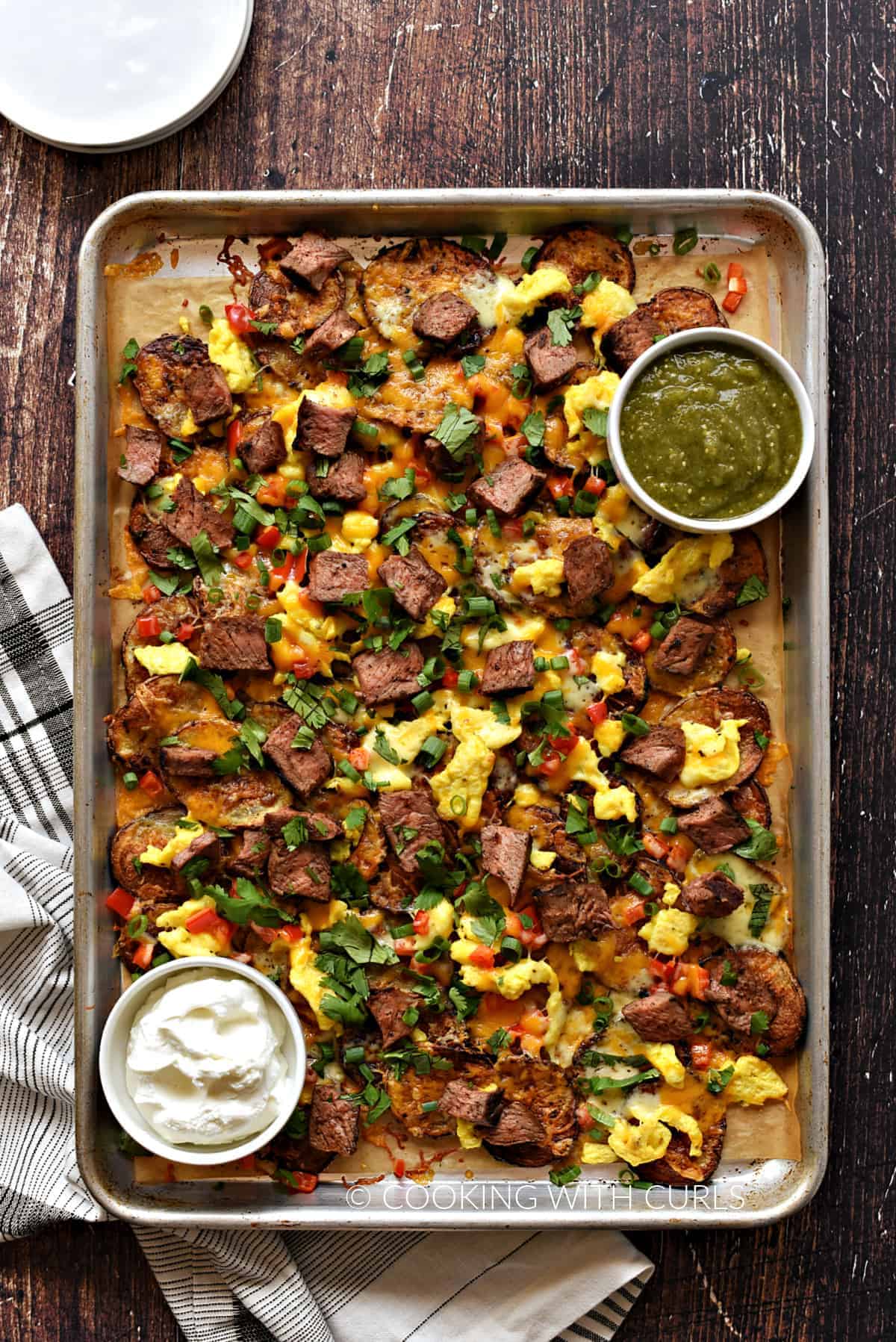 Baked sliced potatoes topped with chopped red pepper and scrambled eggs on a baking sheet with bowls of green salsa and sour cream. 