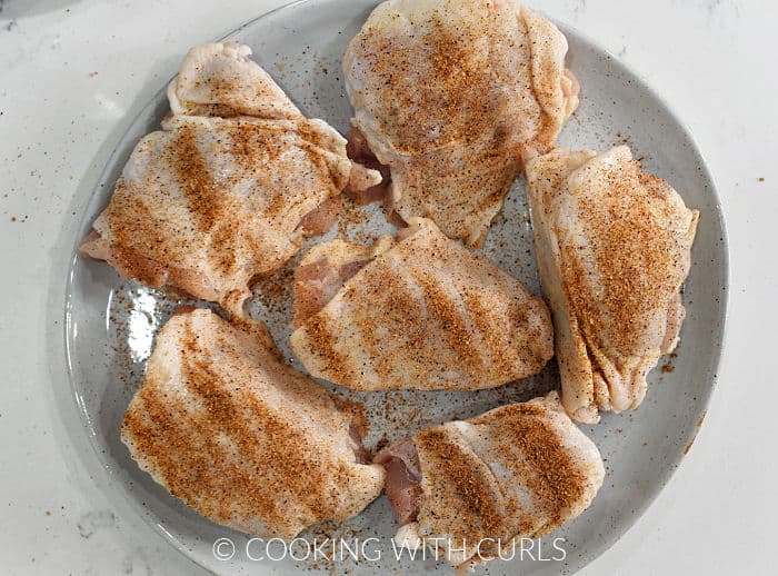 Six chicken thighs on a large plate sprinkled with seasoning.