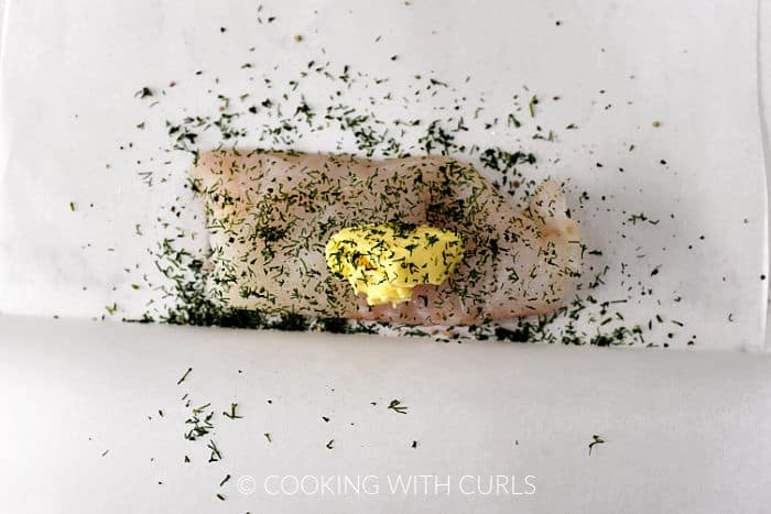 A cod filet on a sheet of parchment paper topped with chopped dill, butter, salt and pepper. 