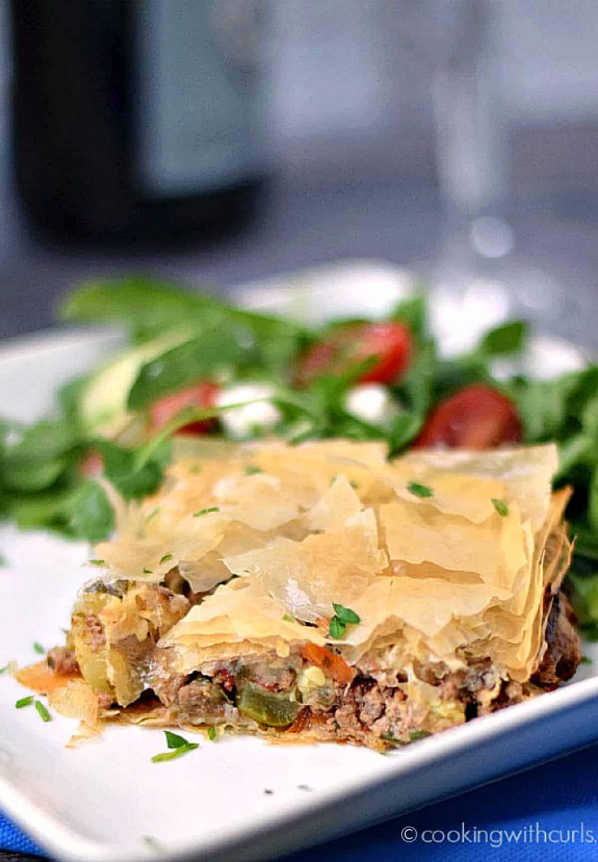 A slice of Greek Summery Meat Pie on a white plate with a Greek salad on the side.