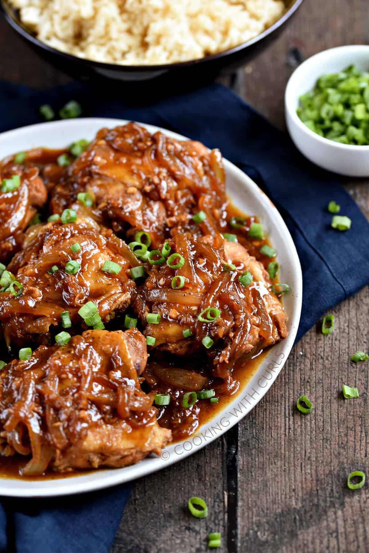 A plate full of chicken thighs covered with a dark brown onion sauce and chopped green onions.
