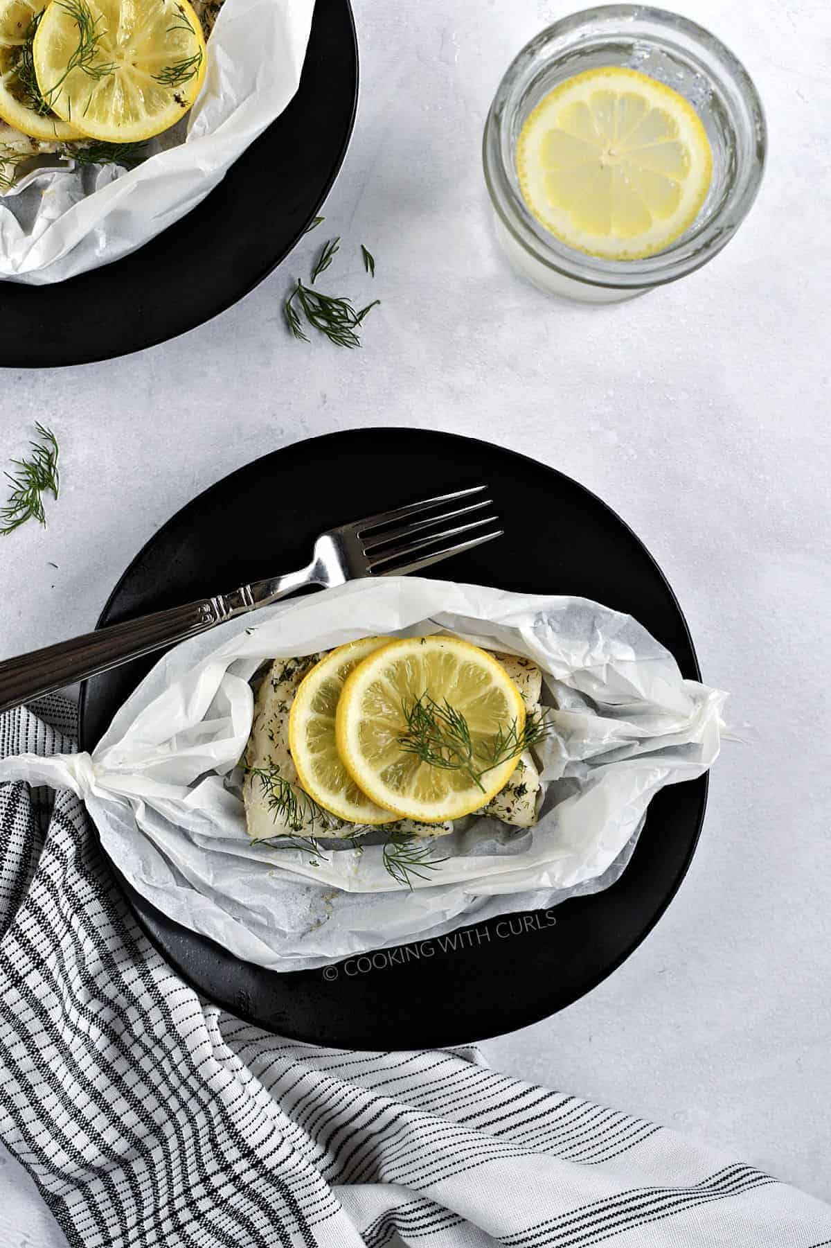 Looking down on two Lemon-Dill Cod fillets baked in Parchment Paper and served on black plates with a glass of lemon water in between the two plates. 