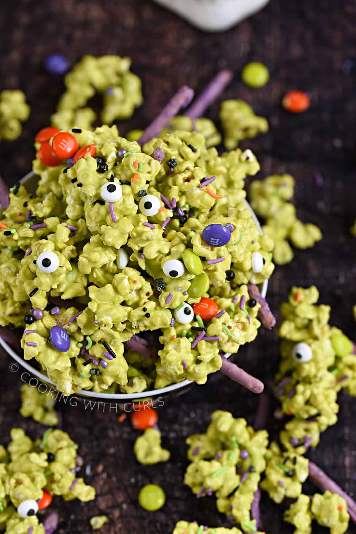 A bowl overflowing with green candy coated popcorn, sprinkles, purple pretzel sticks, and multi-colored candy coated chocolates. 