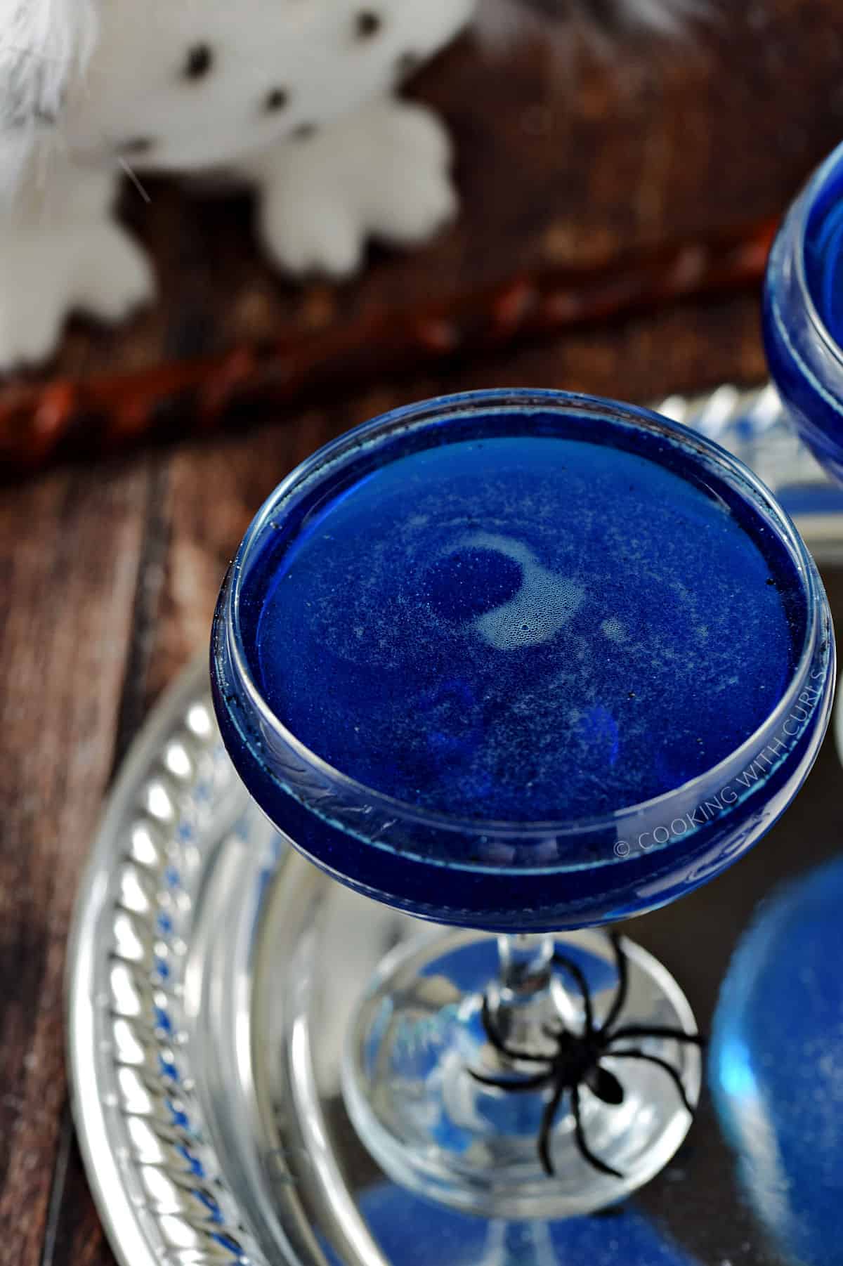 Swirling, shimmering blue drink in a coupe cocktail glass sitting on a silver tray with a spider on the base. 