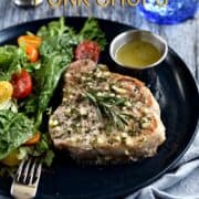Thick, bone-in pork chop topped with garlic-rosemary butter with a green salad and cherry tomatoes on the side with title graphic across the top.
