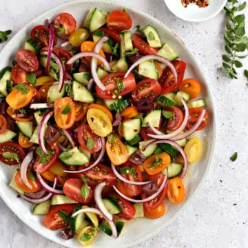Looking down on a large plate with sliced cherry tomatoes, cucumber, kalamata olives, and red onion with fresh oregano leaves and crushed red pepper.