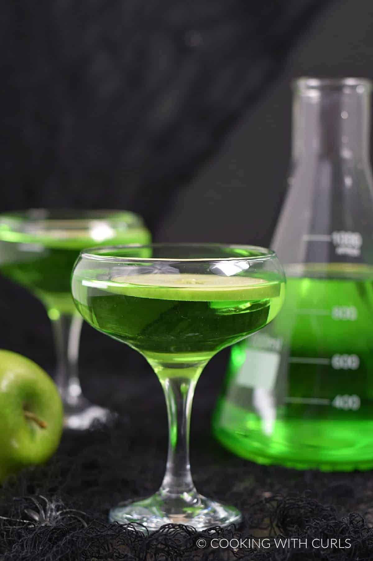 Poisoned Apple Cocktail in two coupe glasses with apple slices, and in a large beaker. 