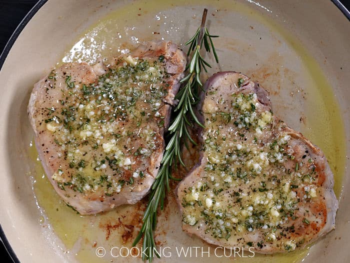 Two pork chops topped with garlic-rosemary butter in a skillet with a fresh rosemary sprig between them. 