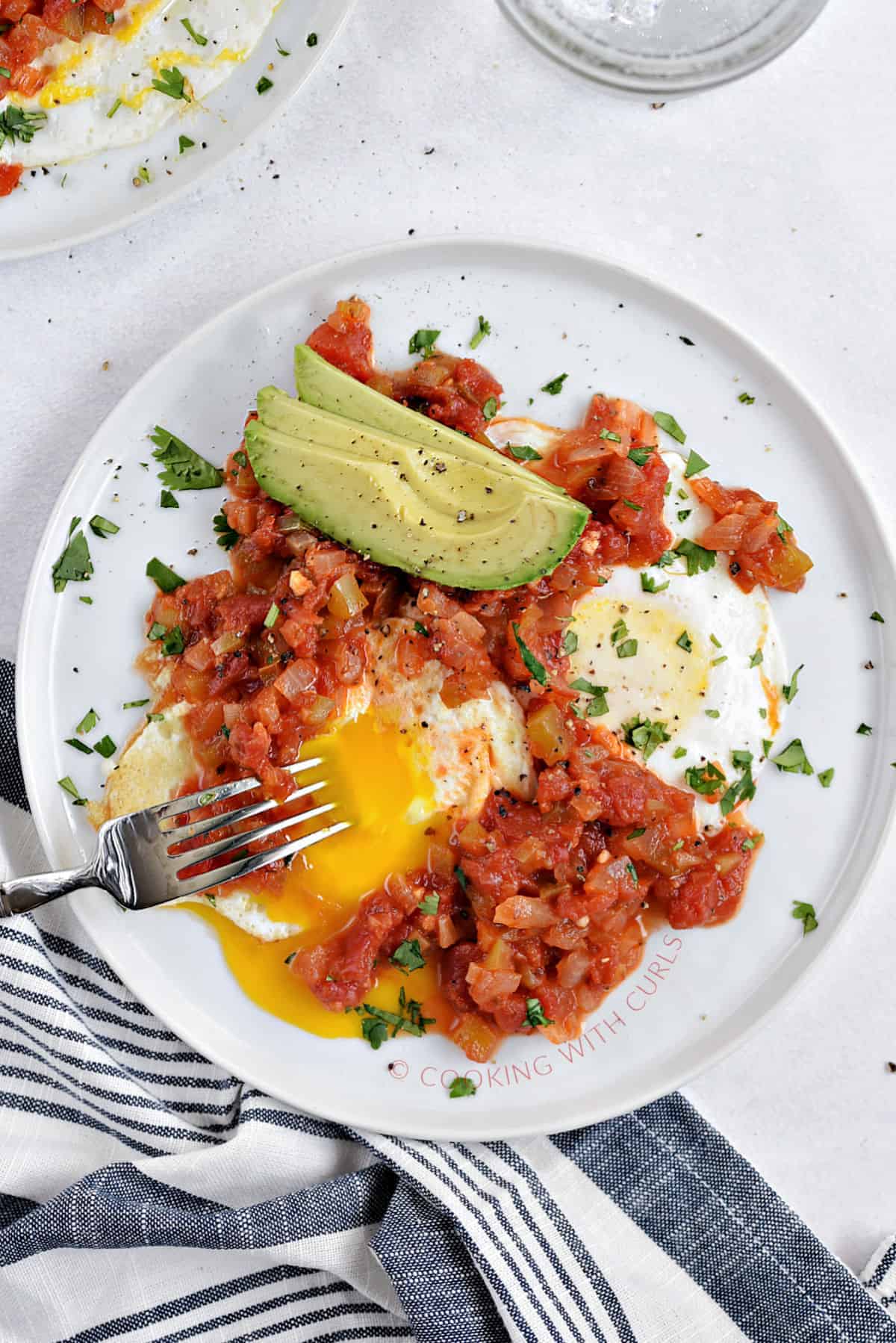 Two fried eggs, one broken open, cooked tomato salsa, and sliced avocado on a small plate.