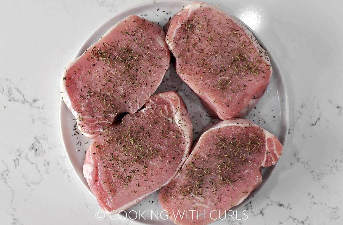 Four pork chops on a plate sprinkled with salt, pepper, and thyme leaves.