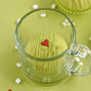 A green chocolate cocoa bomb in a glass mug with a second bomb in the background and tiny marshmallows and red heart candies scattered around the background.