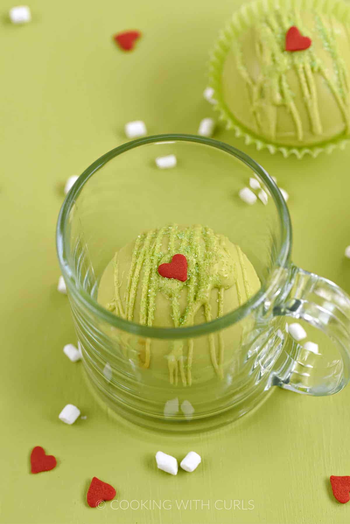 A green chocolate cocoa bomb in a glass mug with a second bomb in the background and tiny marshmallows and red heart candies scattered around the background.