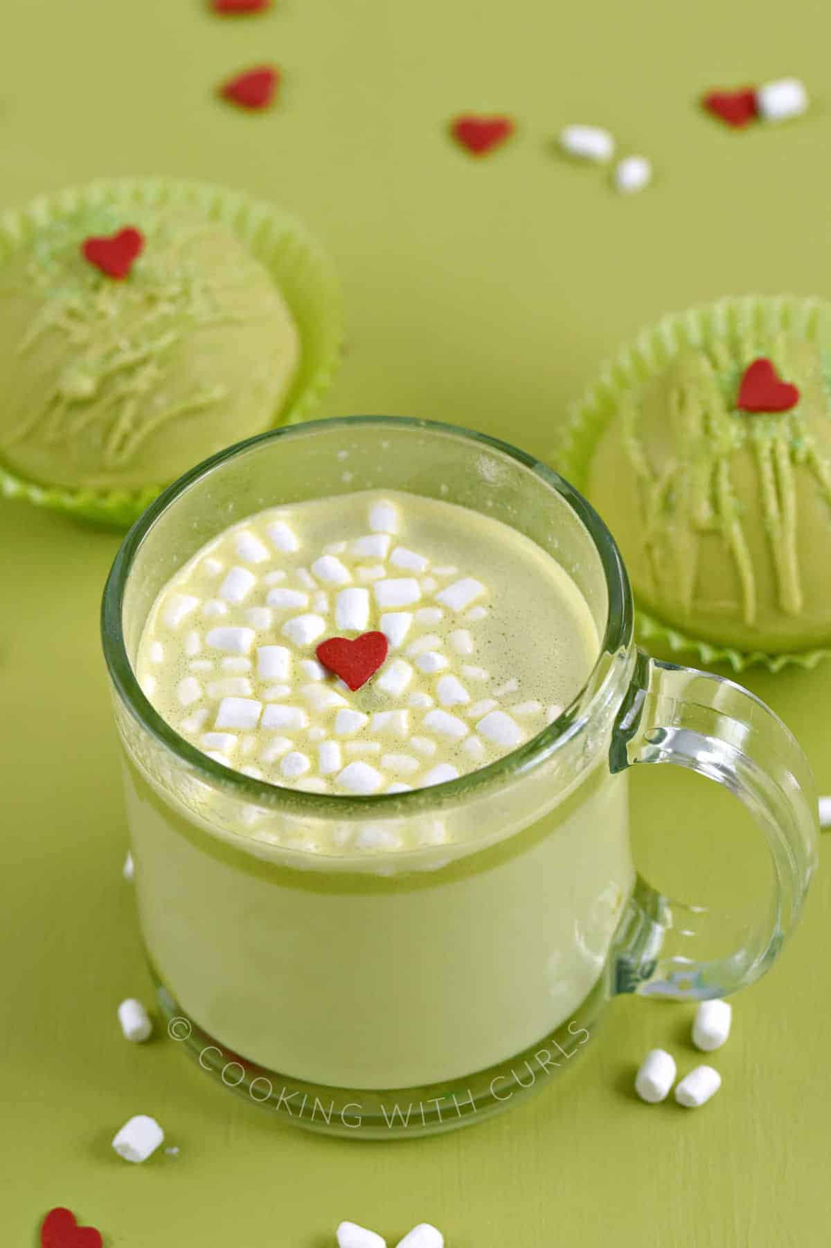 Green hot chocolate in a glass mug topped with mini marshmallows and a red heart candy with two green chocolate bombs in the background. 