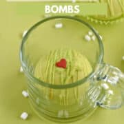 A green chocolate cocoa bomb in a glass mug with a second bomb in the background and tiny marshmallows and red heart candies scattered around the background with title graphic across the top.