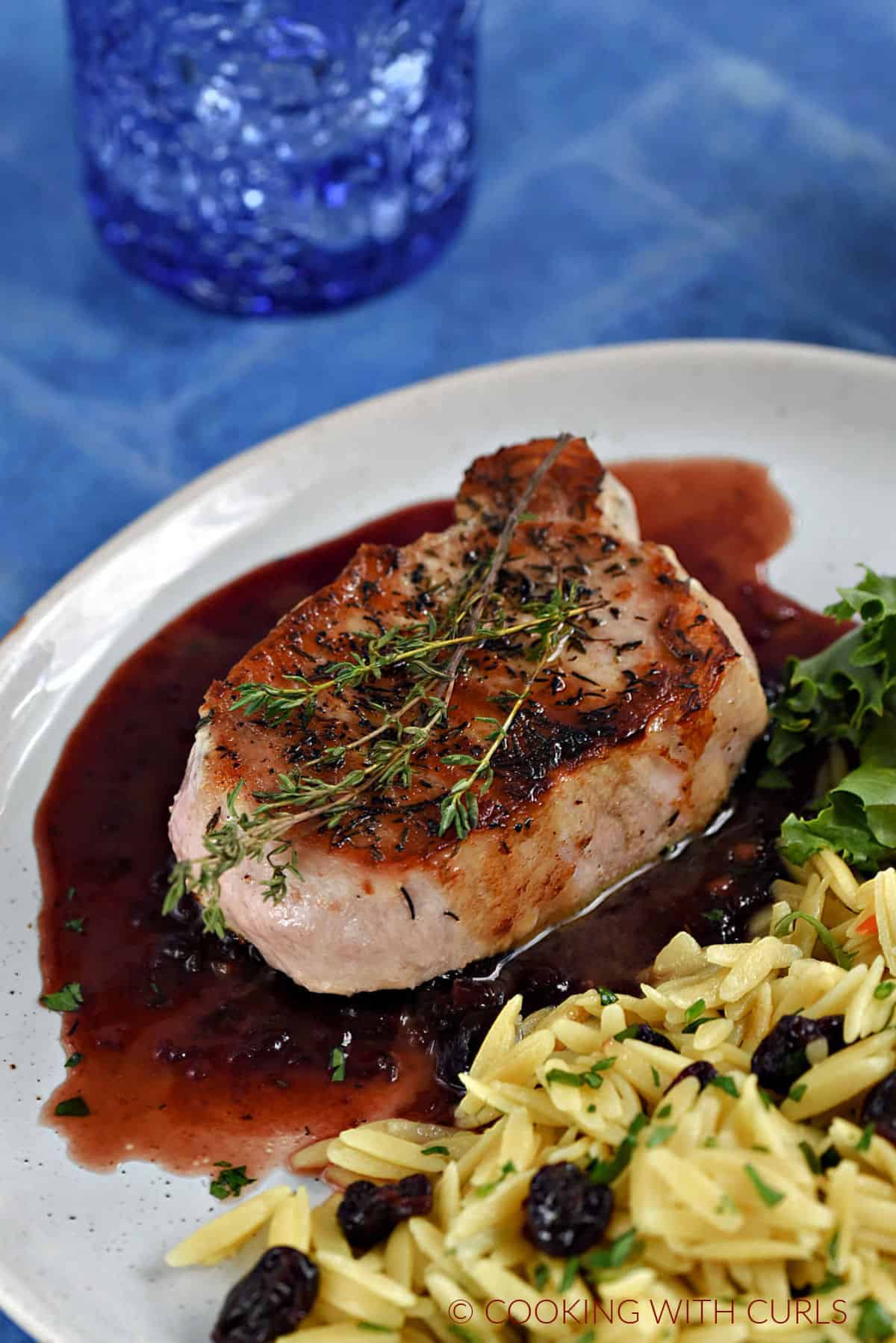 A pork chop sitting on cherry sauce topped with thyme sprigs next to orzo pasta mixed with dried cherries.