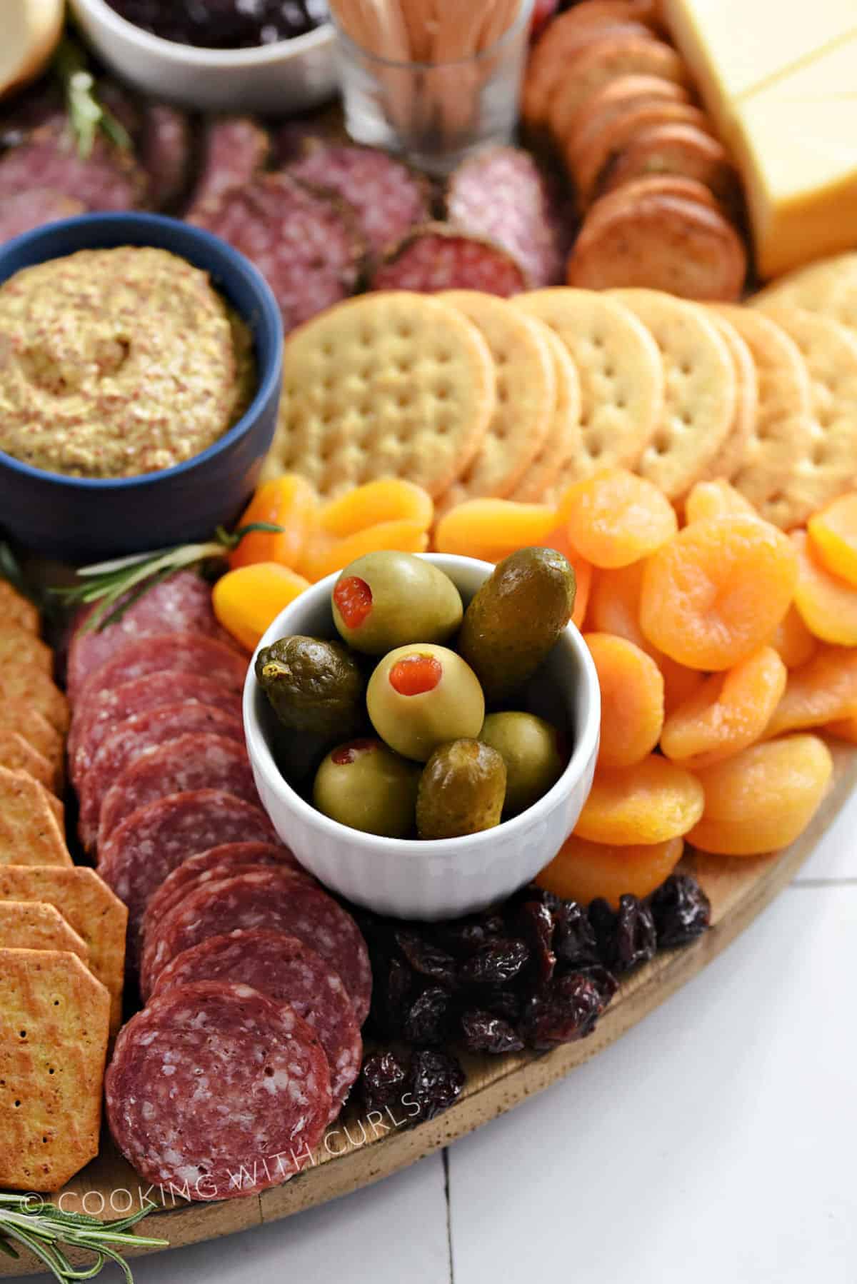 Sliced salami, crackers, green olives in a small bowl, dried cherries and apricots on a large appetizer board.