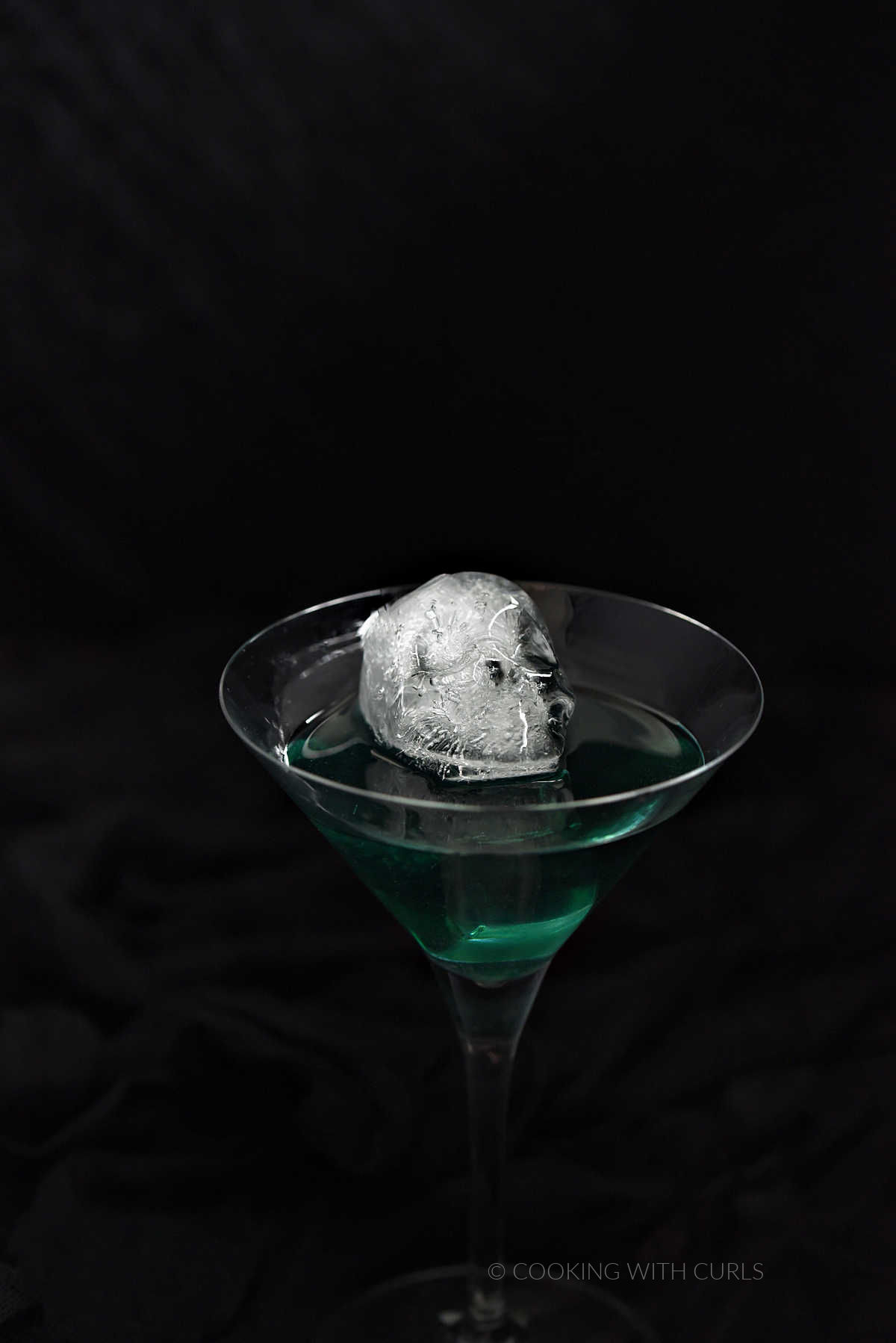 A skull shaped ice cube floating in emerald green liquid in a martini glass.