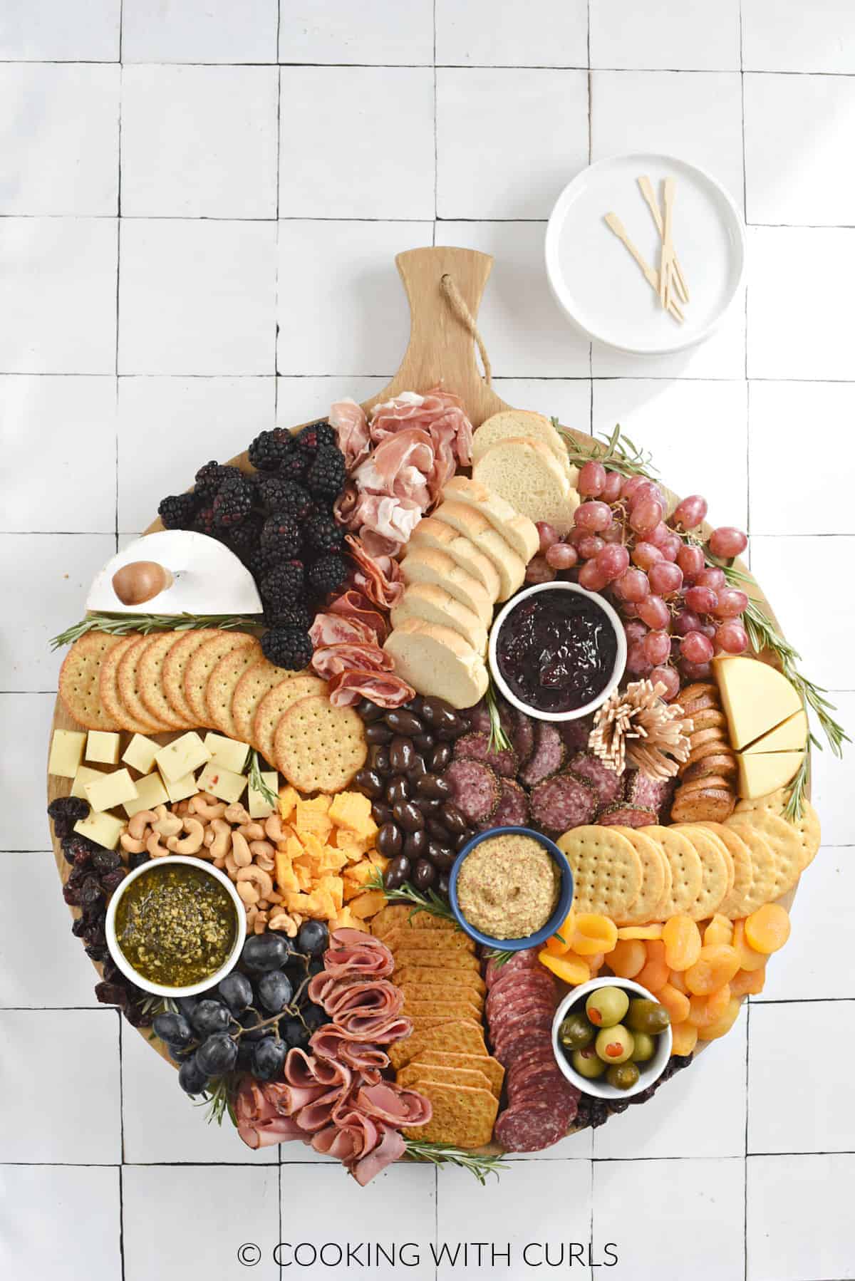 A large round board topped with slices of meats, cheeses, bread, crackers, dried apricots and cherries, grapes, and nuts.