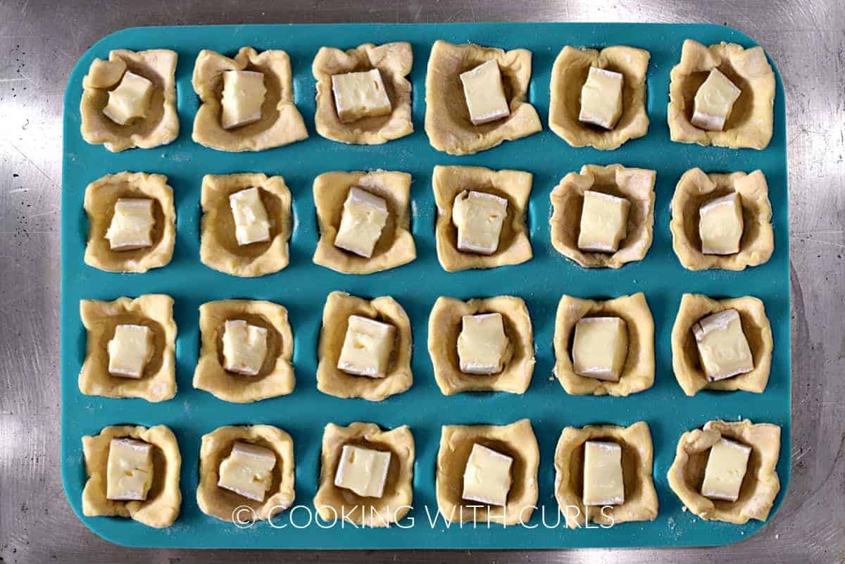 A mini muffin pan sitting on a baking sheet with twenty four puff pastry squares with brie cubes. 