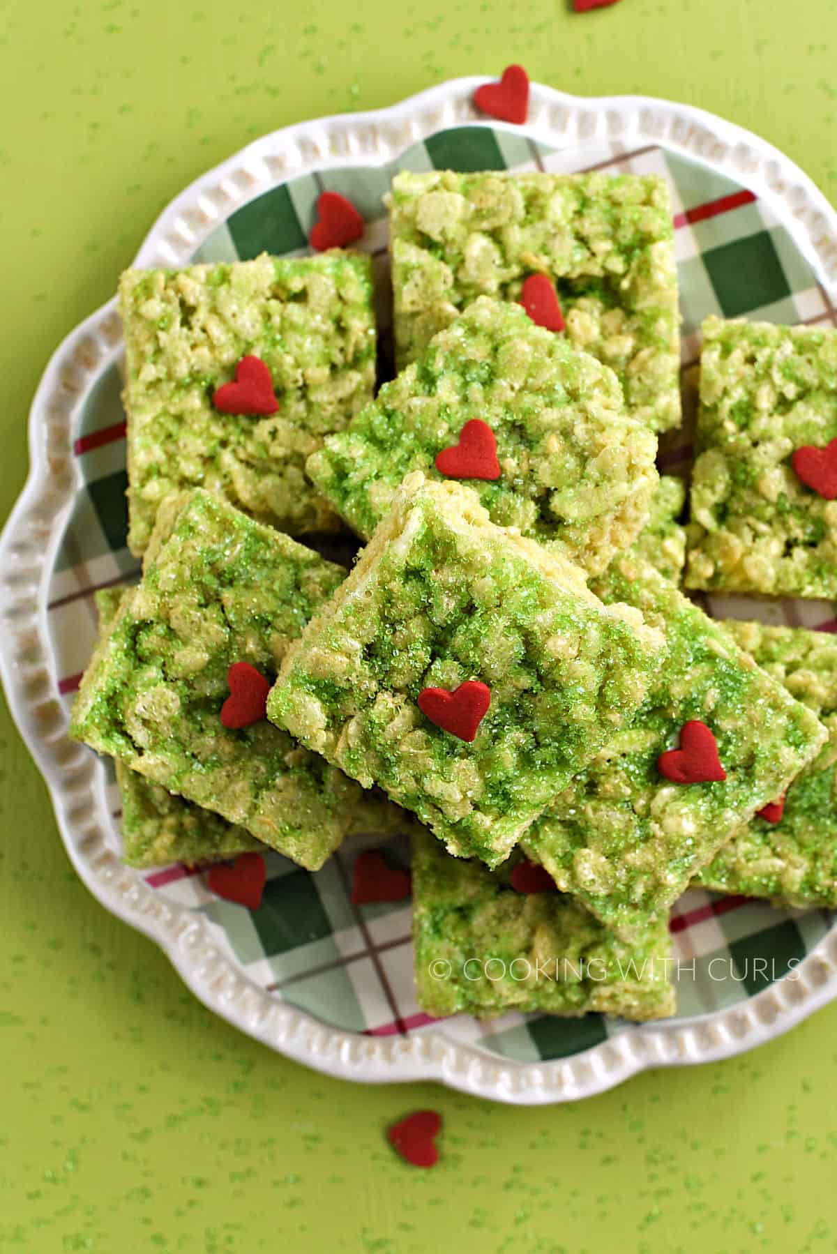 Looking down on a pile of Grinch green rice krispie treats topped with green sugar and jumbo red hearts on a plate.