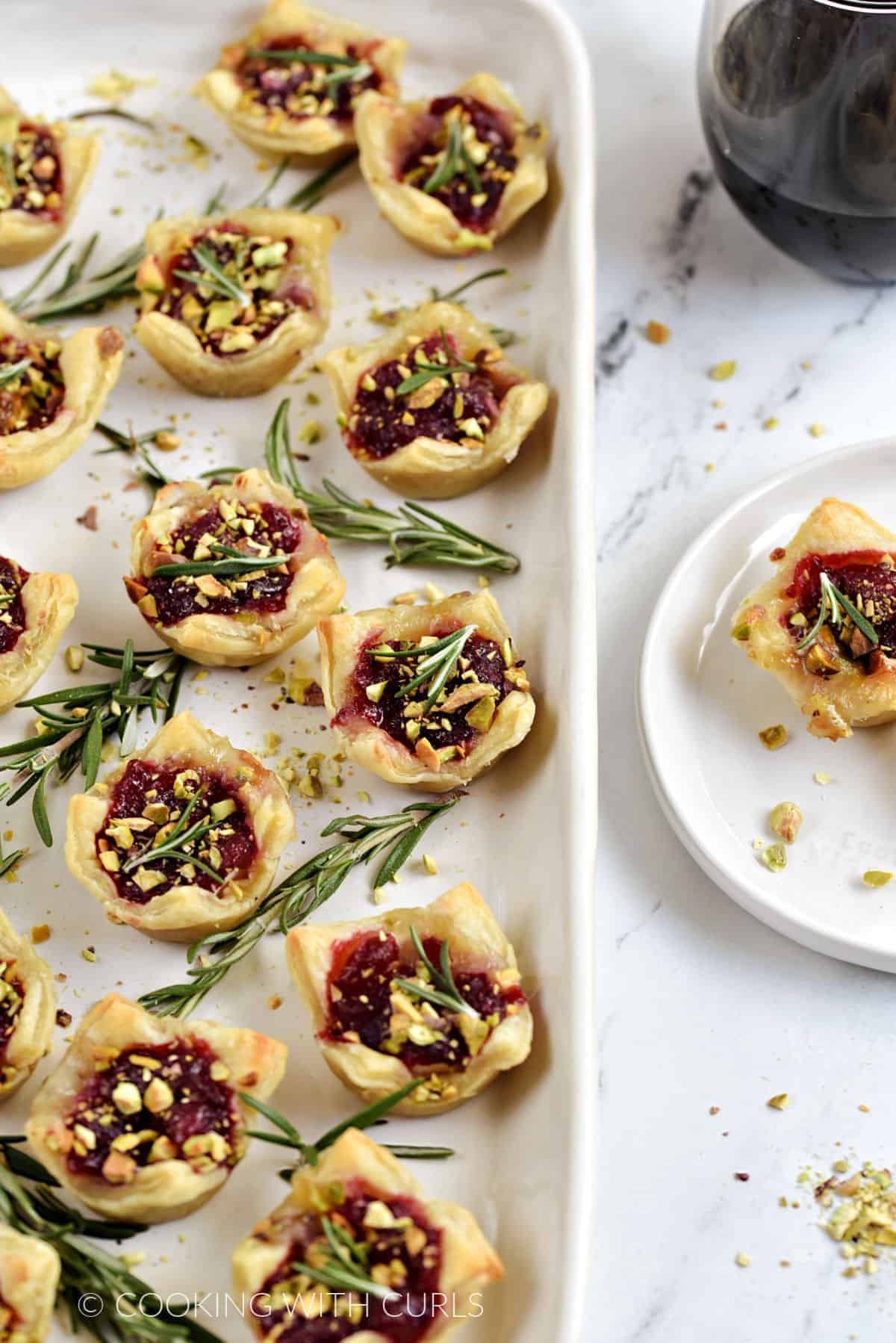Easy, baked Cranberry Brie Bites with sprigs of fresh rosemary on a serving platter with one on a small plate on the right hand side. 