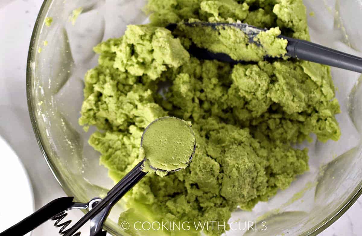 Green cookie dough mixed in a large bowl with a cookie scoop filled with dough in the bottom left corner. 