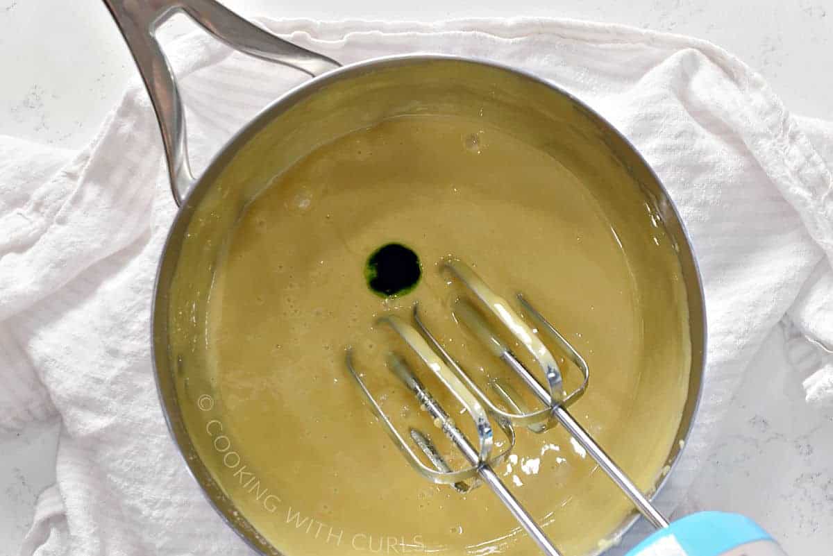 Green food coloring added to the cooled fudge in a saucepan with beaters resting on the side.