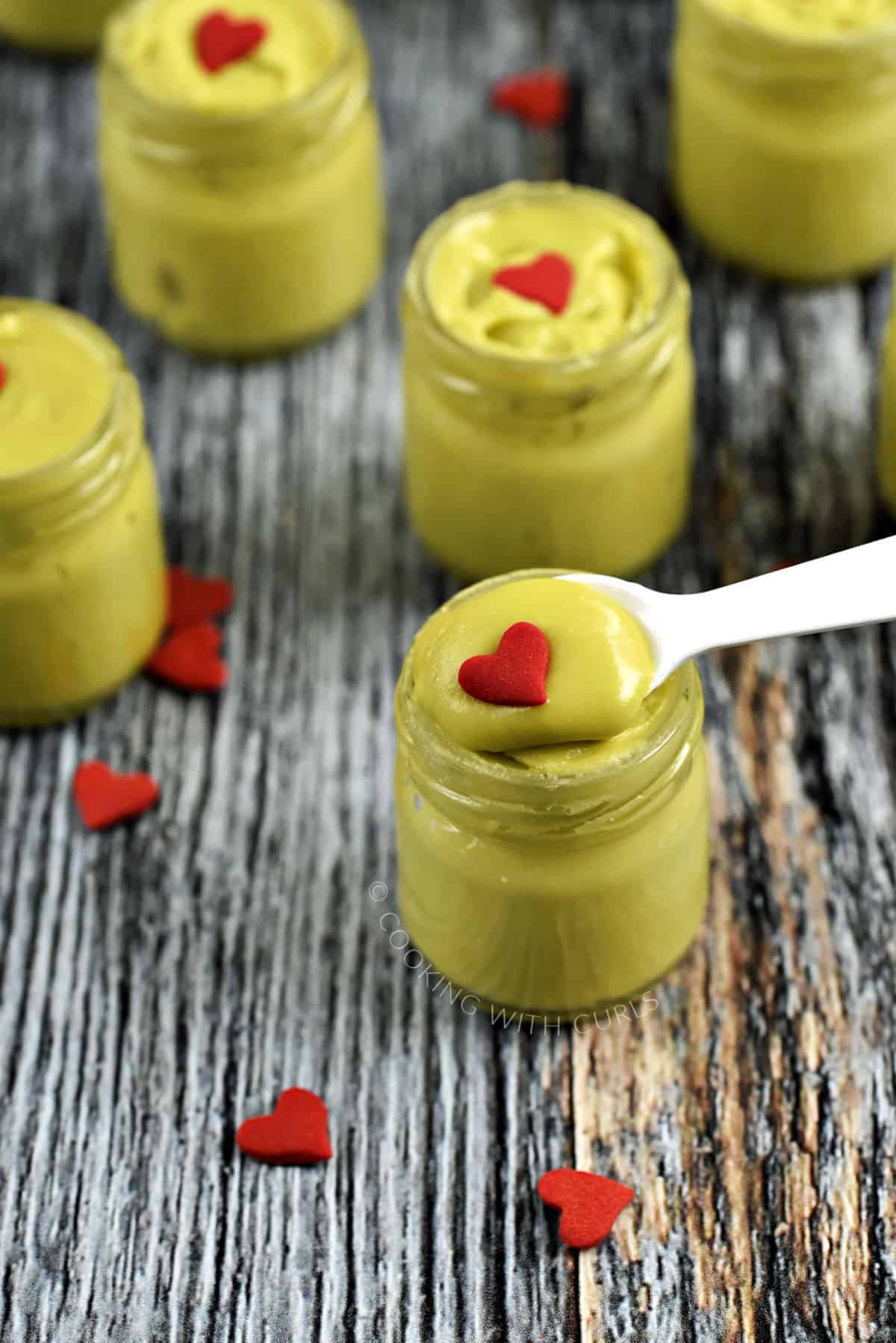 A small white spoon holding green fudge in a jar topped with a red heart with four more jars in the background.
