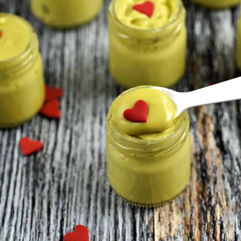 A small white spoon holding green fudge in a jar topped with a red heart with three more jars in the background.