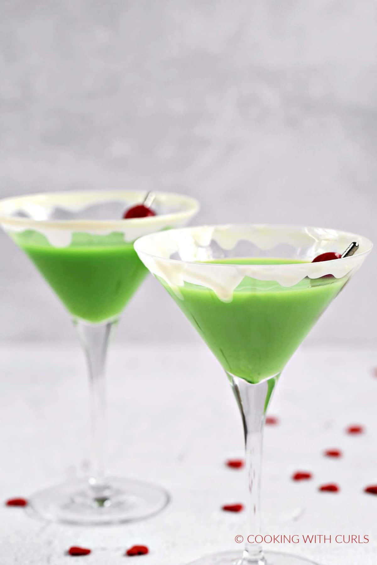 Two green grinch martinis with white drips around the rim and cherry on a cocktail pick surrounded by tiny red heart candies.