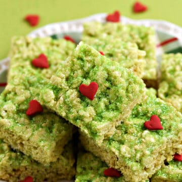 A pile of green rice krispie treats with red hearts piled on a plate .
