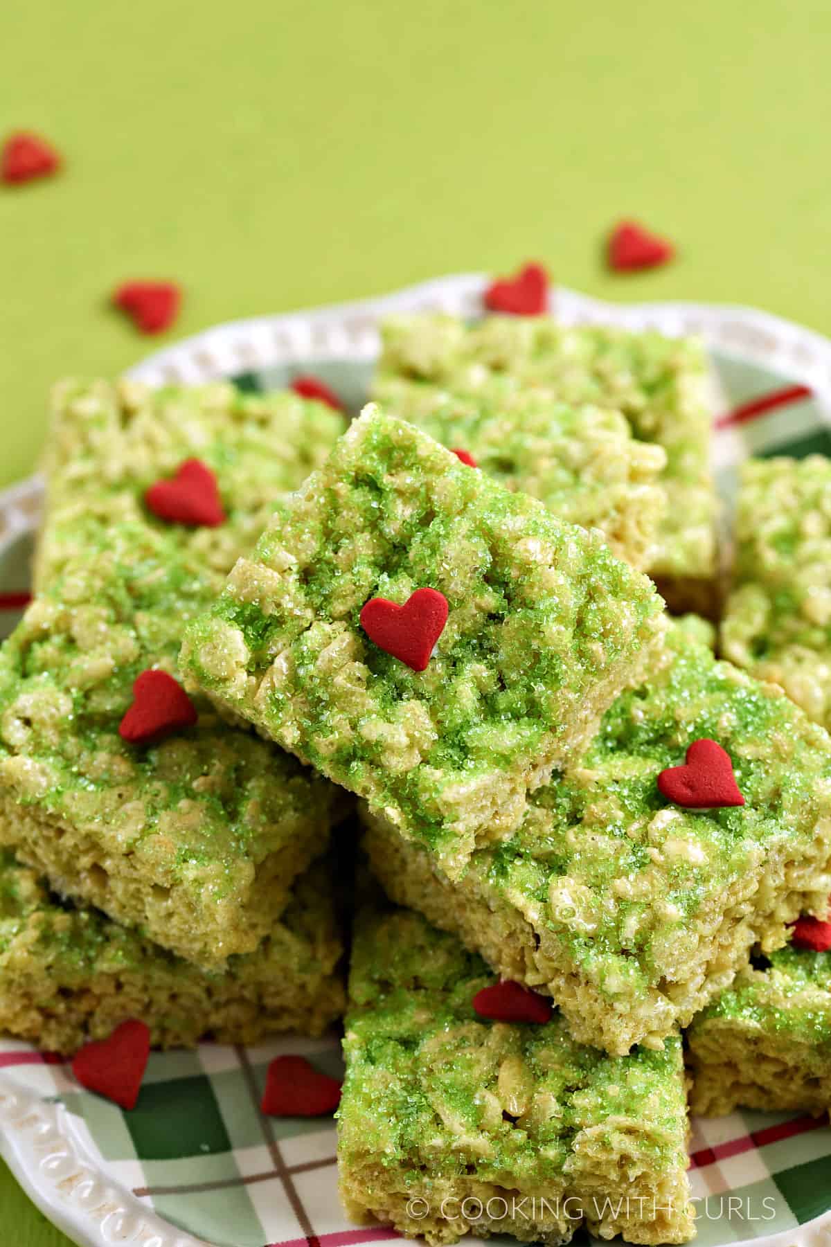 A pile of Grinch green rice krispie treats topped with green sugar and jumbo red hearts on a plate.
