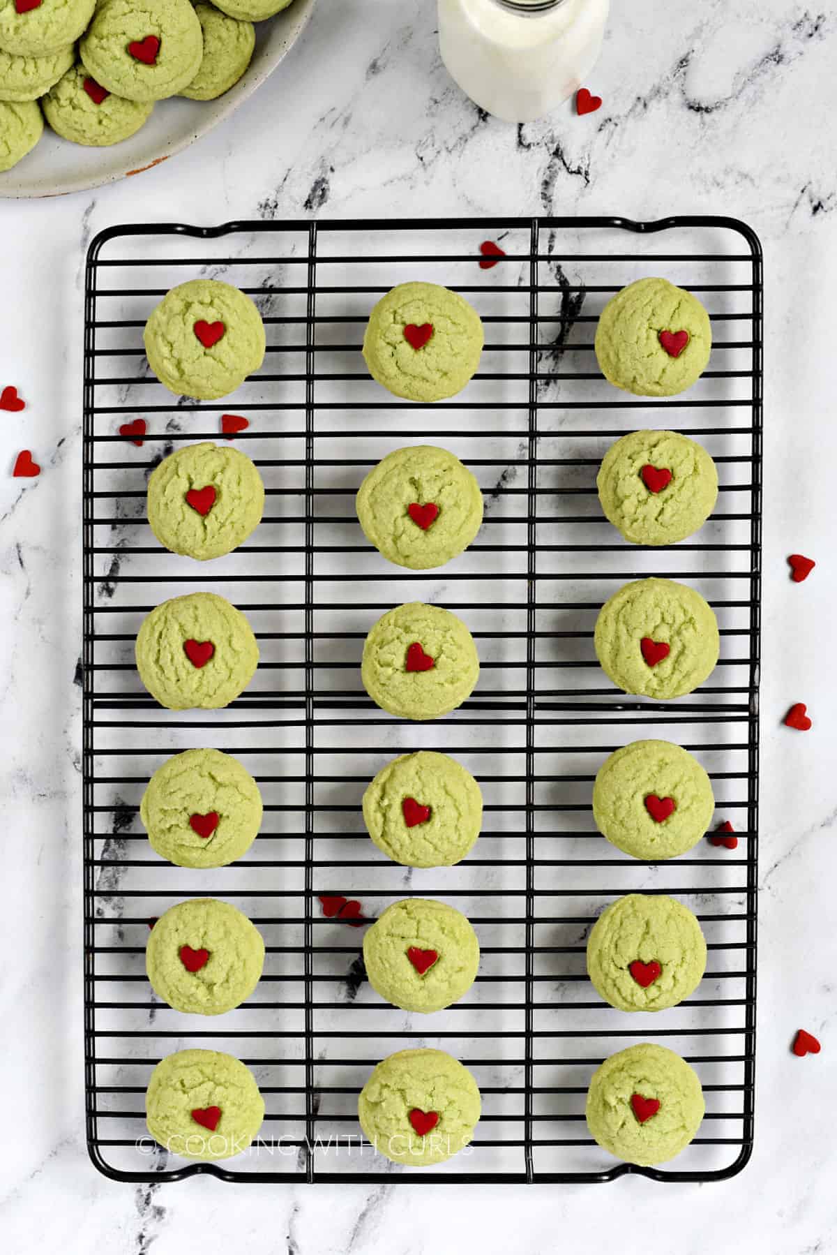 Looking down on 18 Grinch Sugar Cookies on a wire cooling rack with more on a plate in the background.